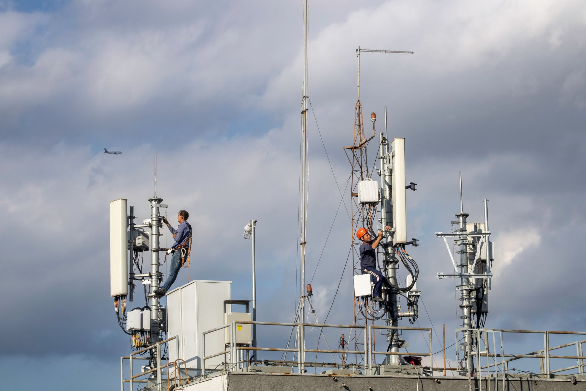Two workers repairing a cellular tower against a cloudy sky,