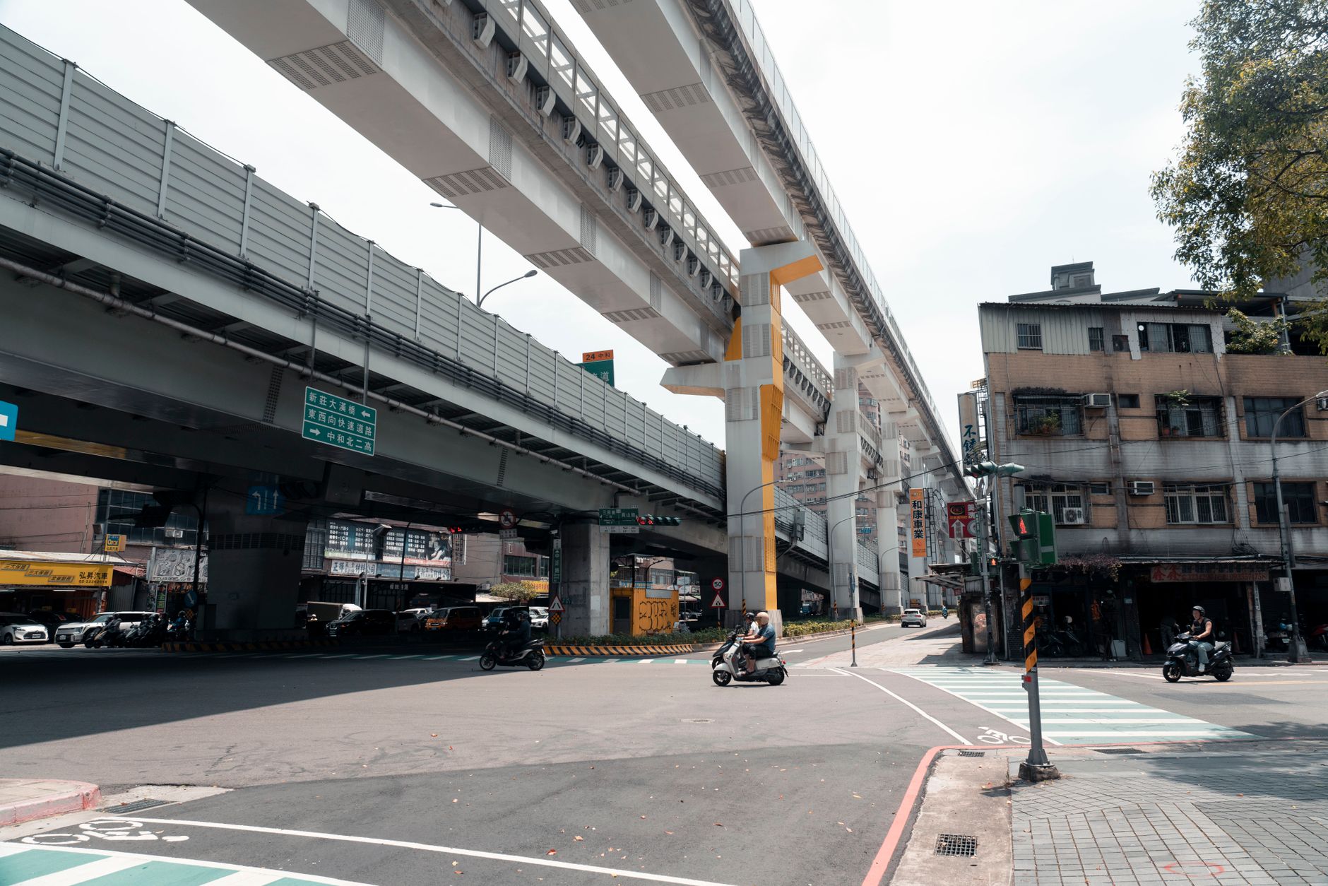 Motorbikes navigate an urban intersection beneath towering elevated train tracks in bustling New Taipei City.
