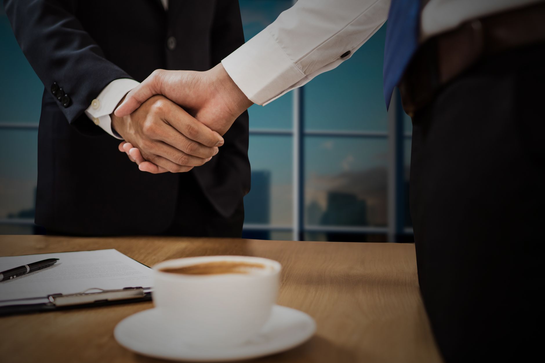 Two professionals shaking hands across a desk with cityscape
