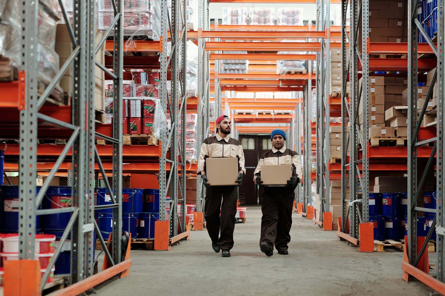 Two workers carrying boxes walk between metal shelves in a s