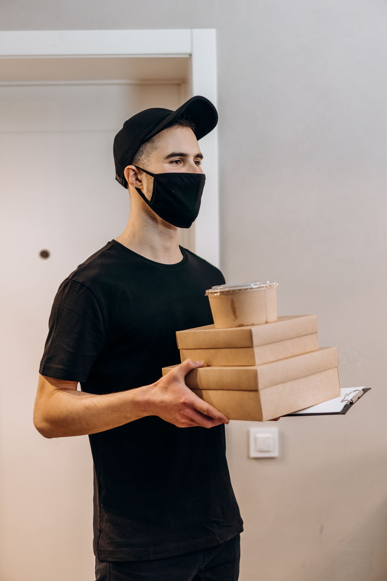 Young delivery man in black mask holding food boxes and clipboard indoors.
