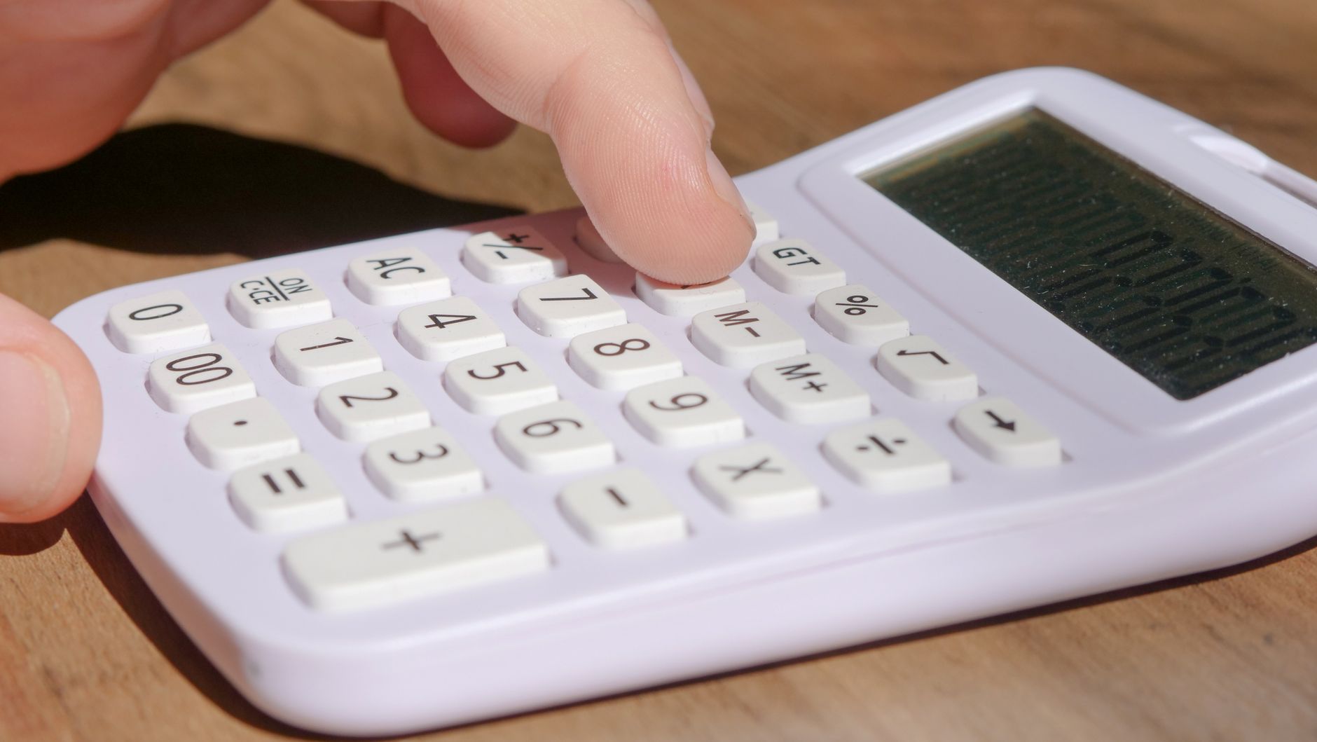 A hand pressing keys on a white calculator placed on a woode