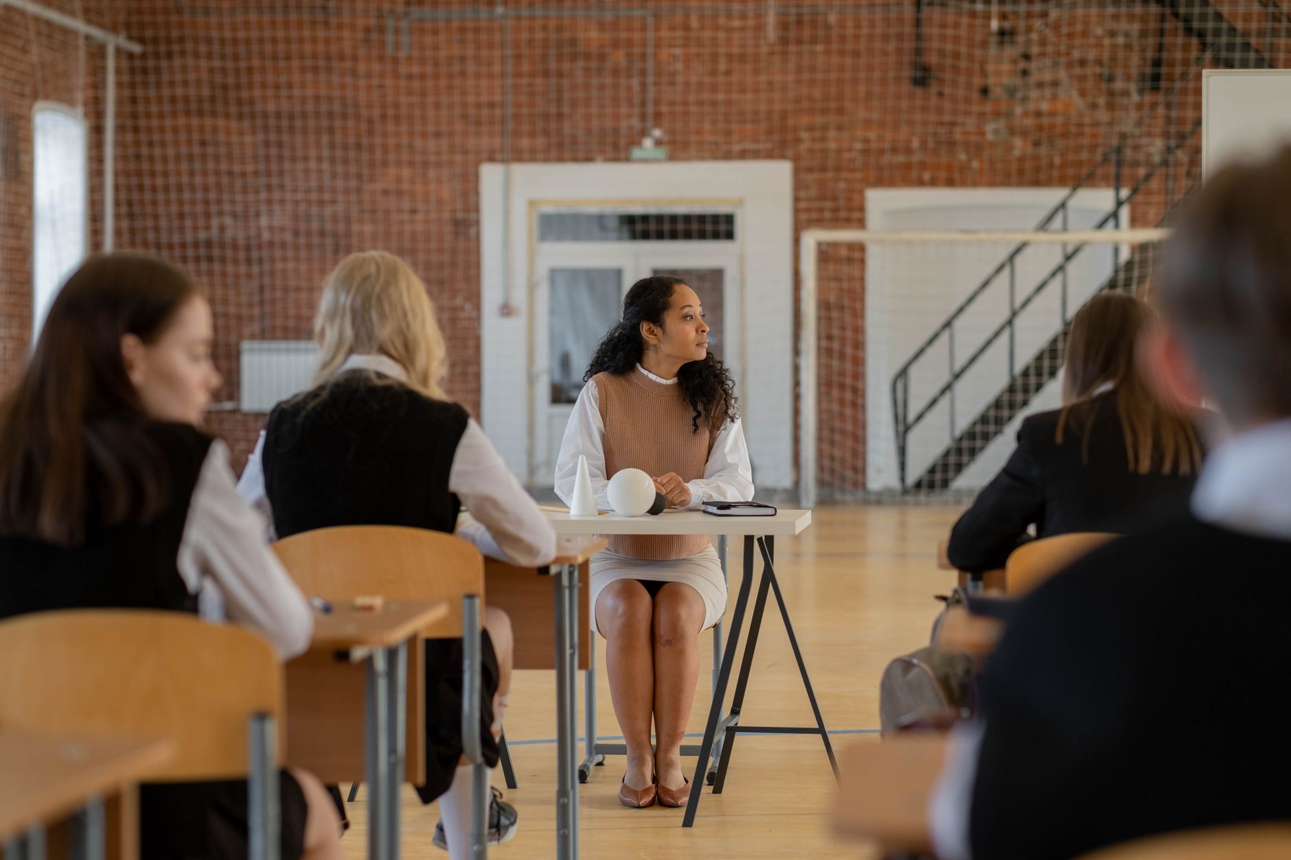 Teacher sitting at desk during class in a school gymnasium.