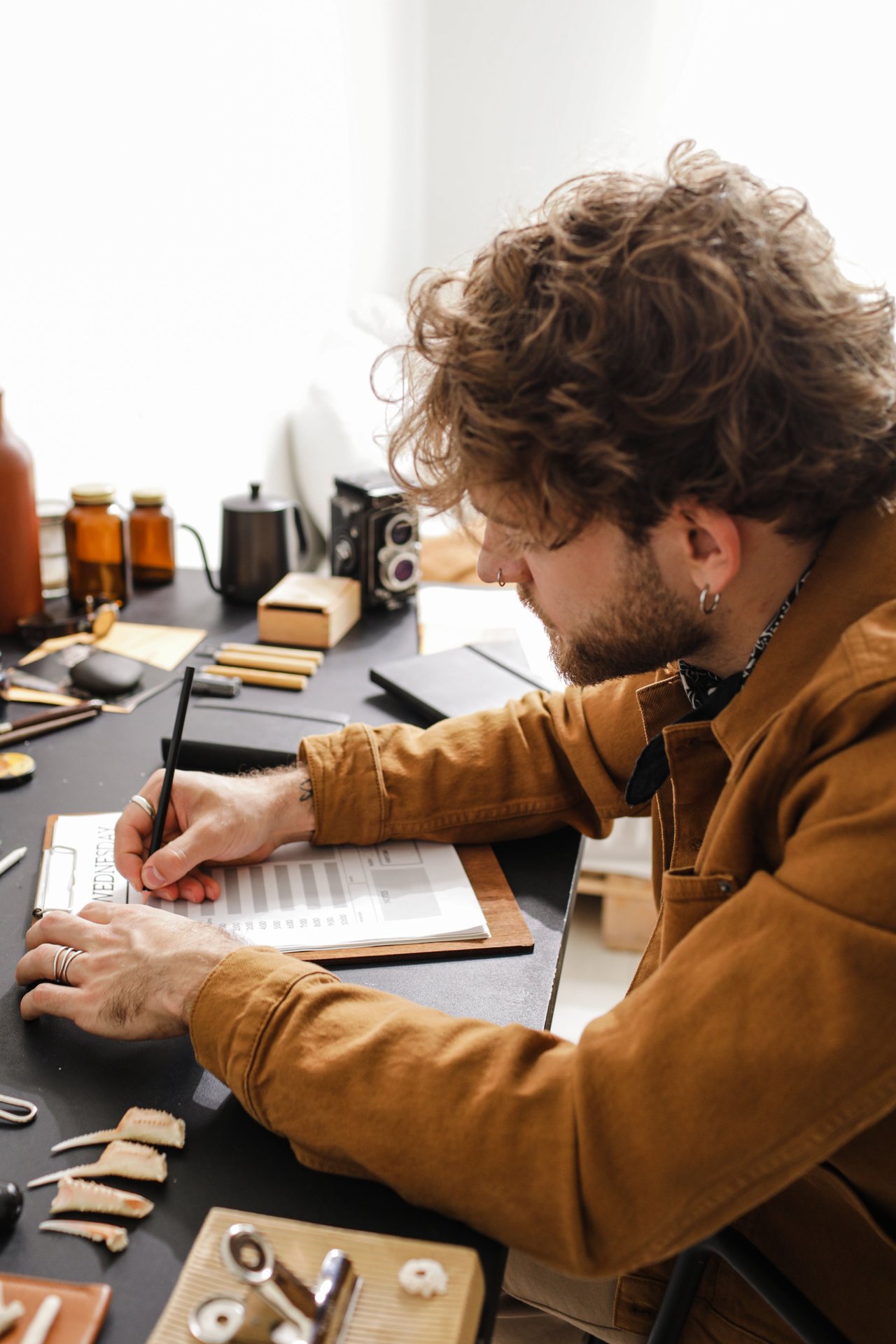 A man in a casual shirt makes notes at a cluttered desk in an artistic workspace.