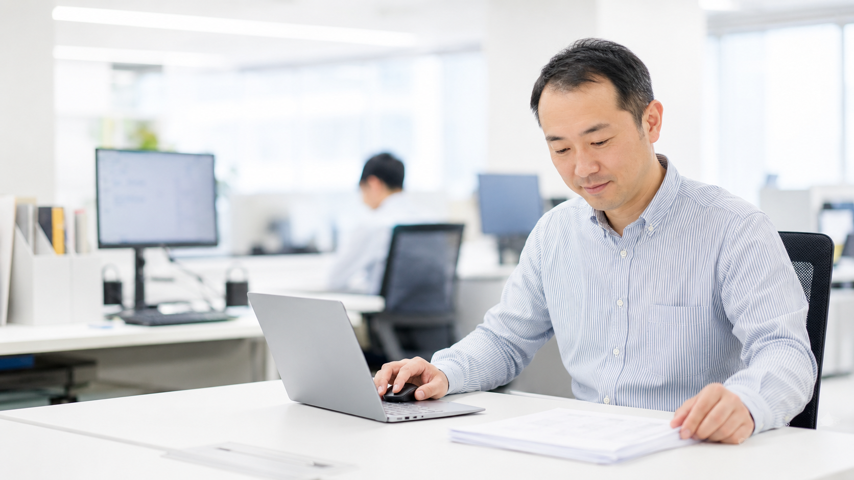 オフィスの自席で納品書とノートパソコンを見比べる日本人の会社員