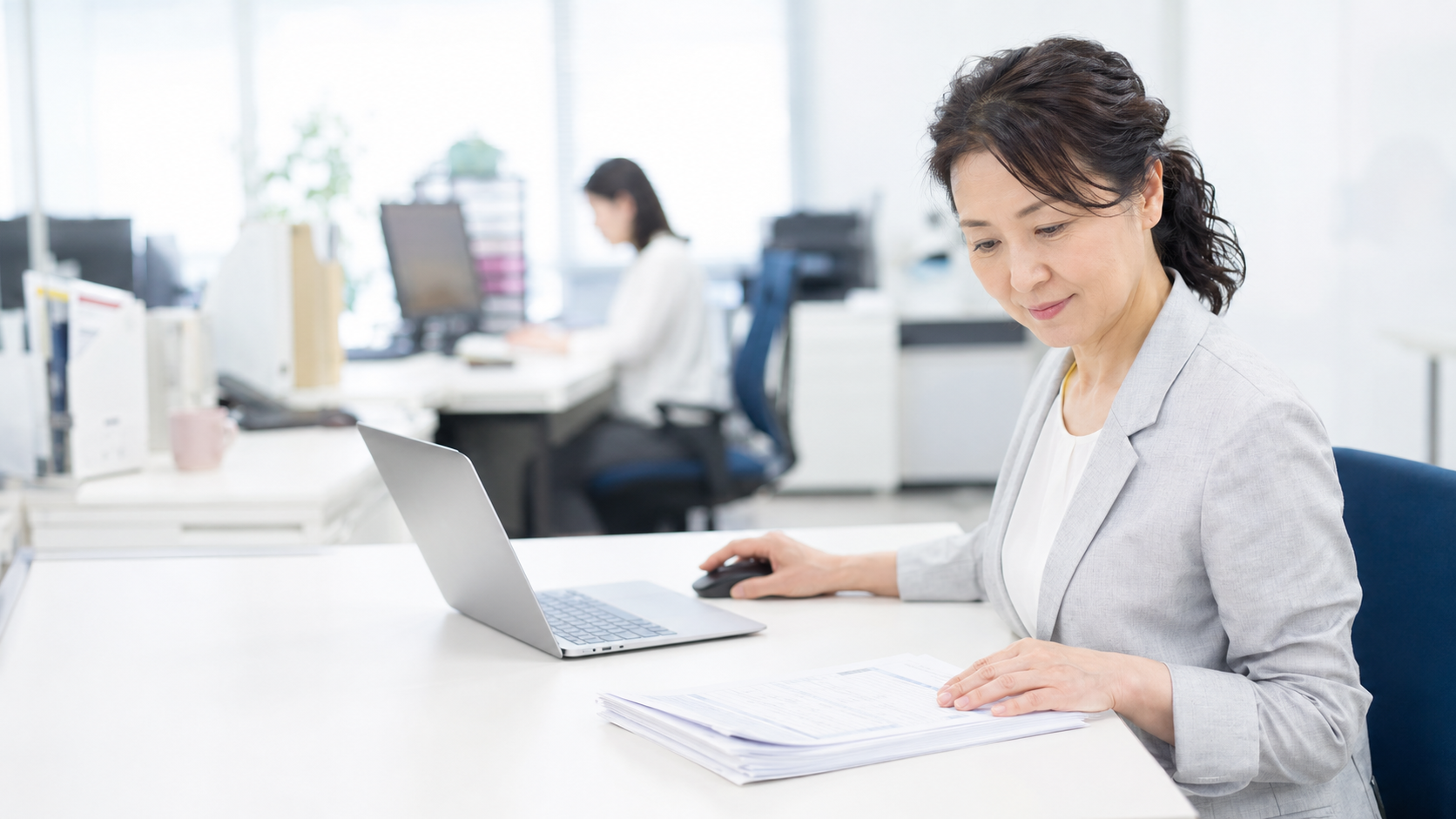 オフィスの自席で納品書とノートパソコンを見比べる日本人の会社員