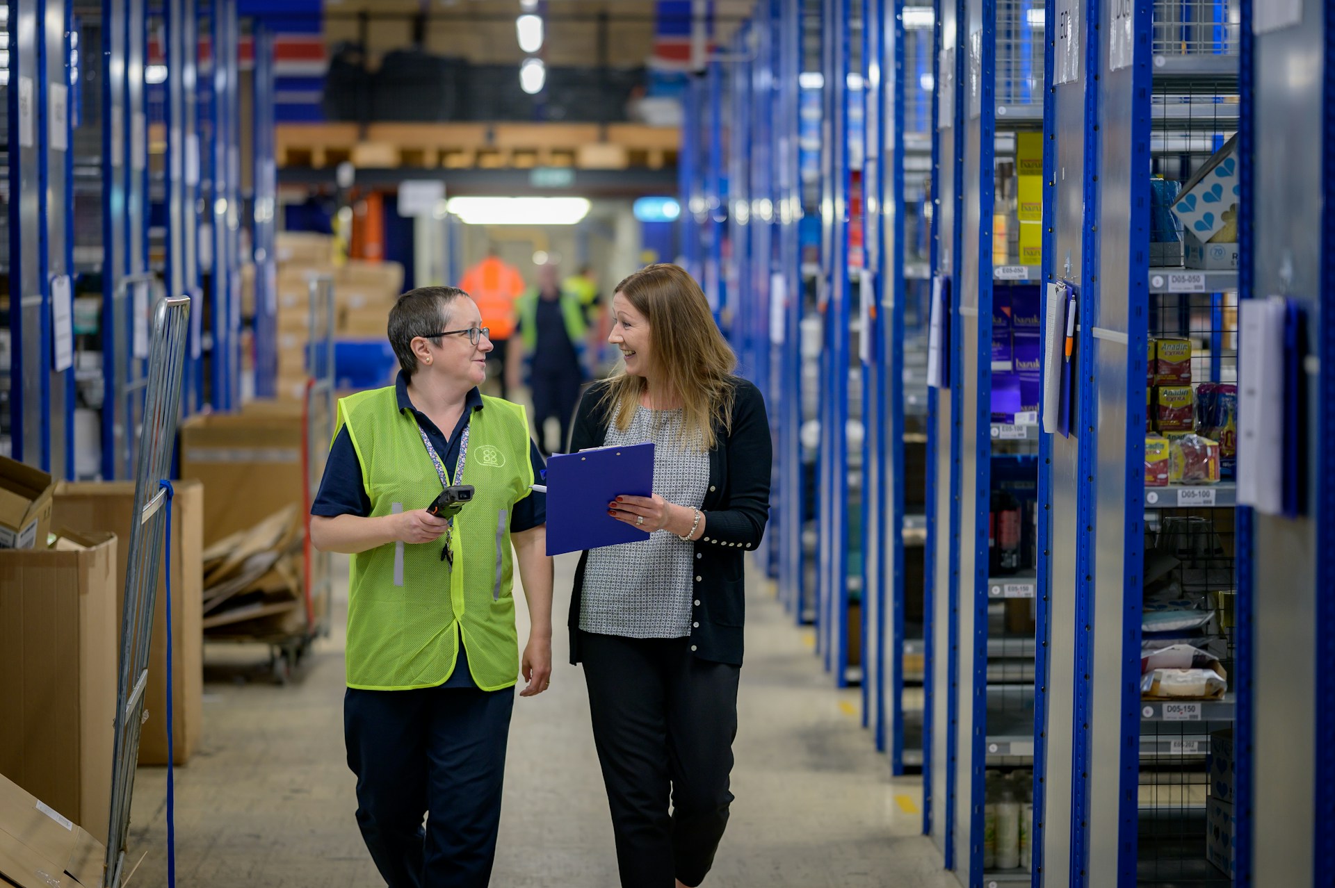 Two women walking through a warehouse aisle with blue shelvi