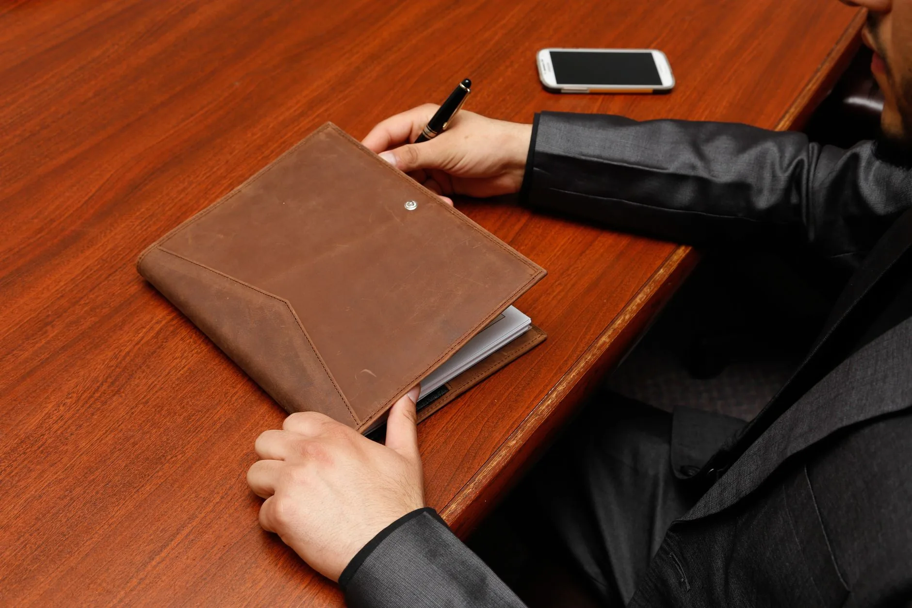 Male executive writing notes during a business meeting using a leather notebook.