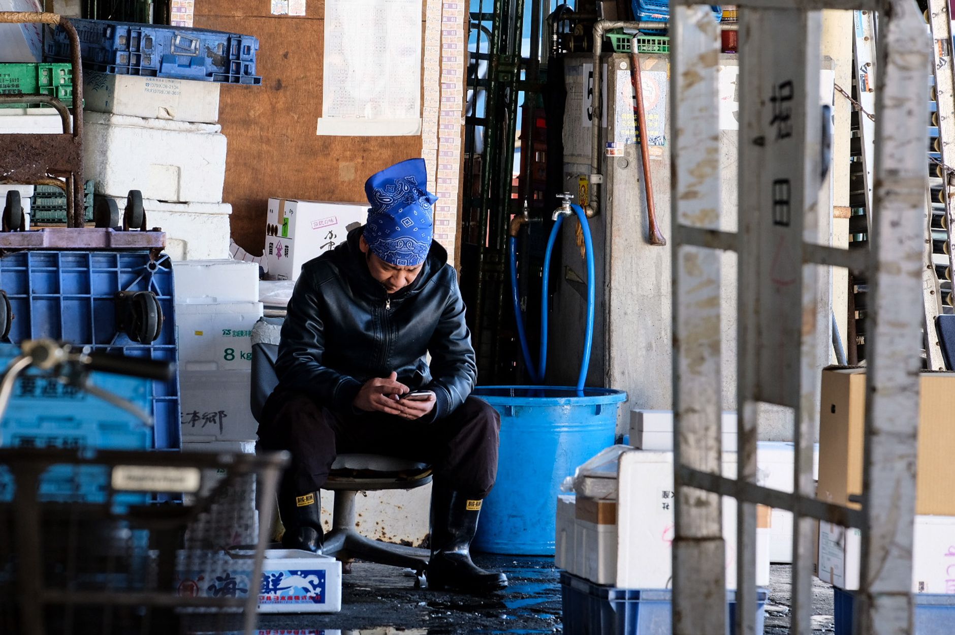 Warehouse worker sitting on a chair, looking at a smartphone, surrounded by boxes and containers.
