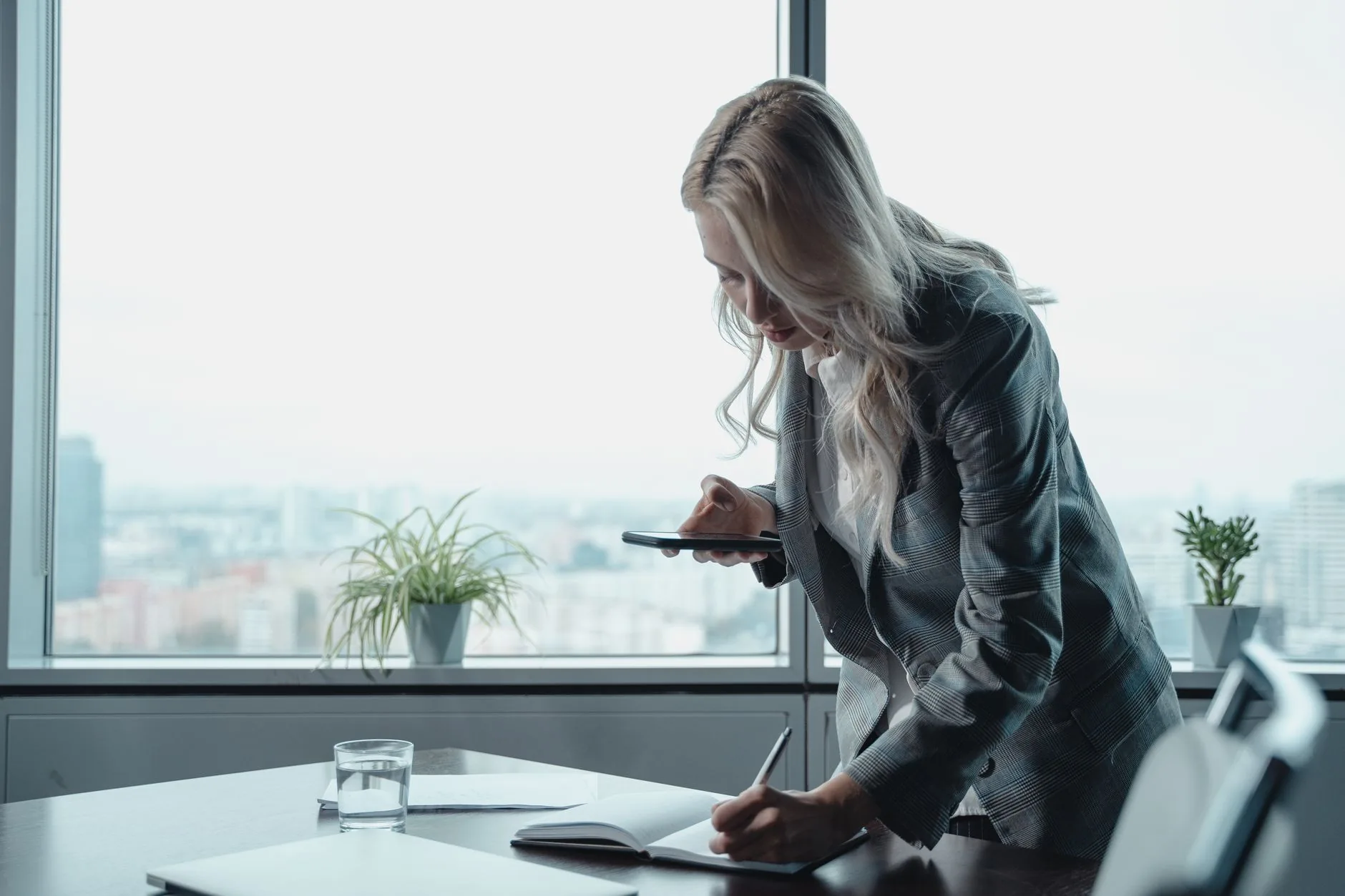 Businesswoman with blonde hair writing notes with a smartphone in a modern office setting.
