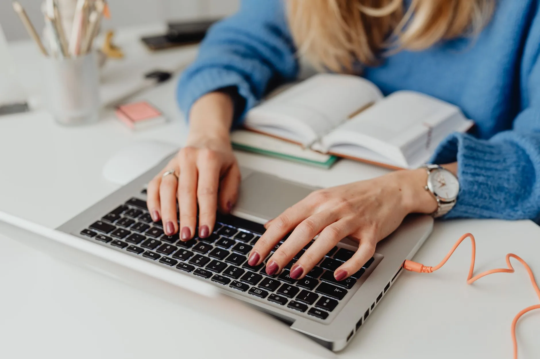 Close-up of hands on laptop keyboard, alongside an open book and stationery tools, in an indoor setting.