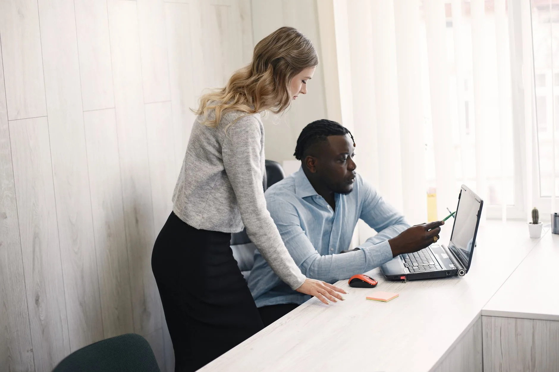 Two coworkers engaged in discussion while using a laptop in a modern office setting.