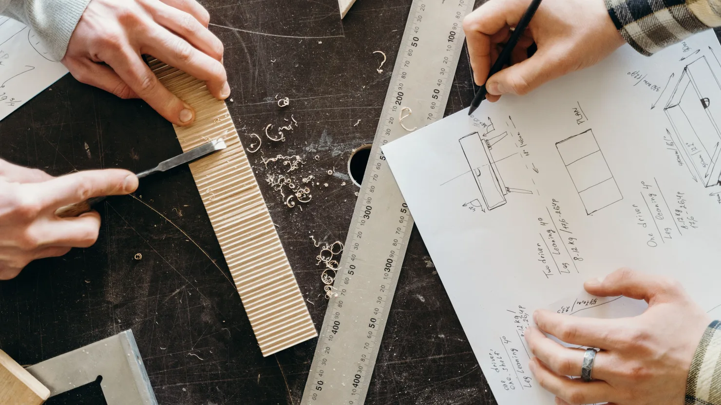 Close-up of hands designing woodworking projects on paper with rulers and wood shavings.