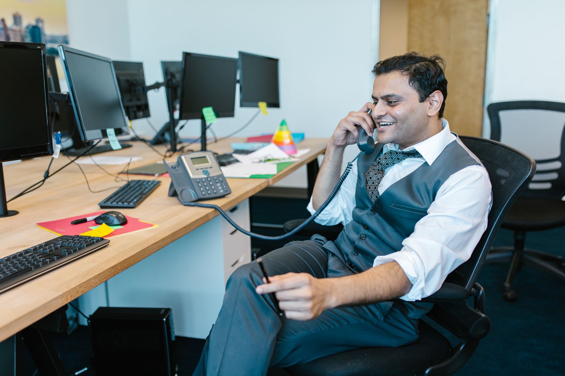 A businessman in formal attire talks on the phone while seated at a computer desk in a modern office.