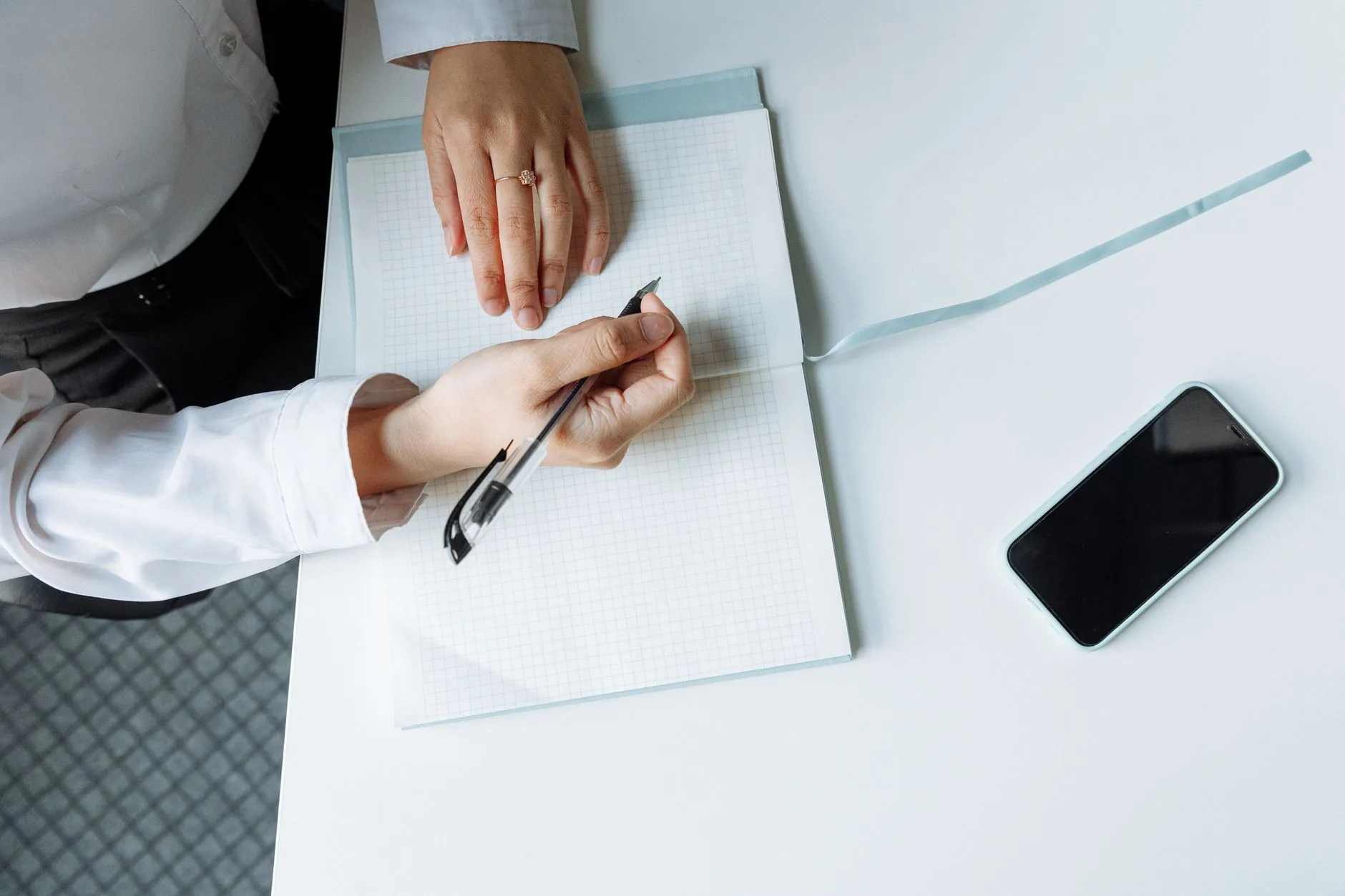 Close-up of hands writing in a notebook with a smartphone on a white desk.