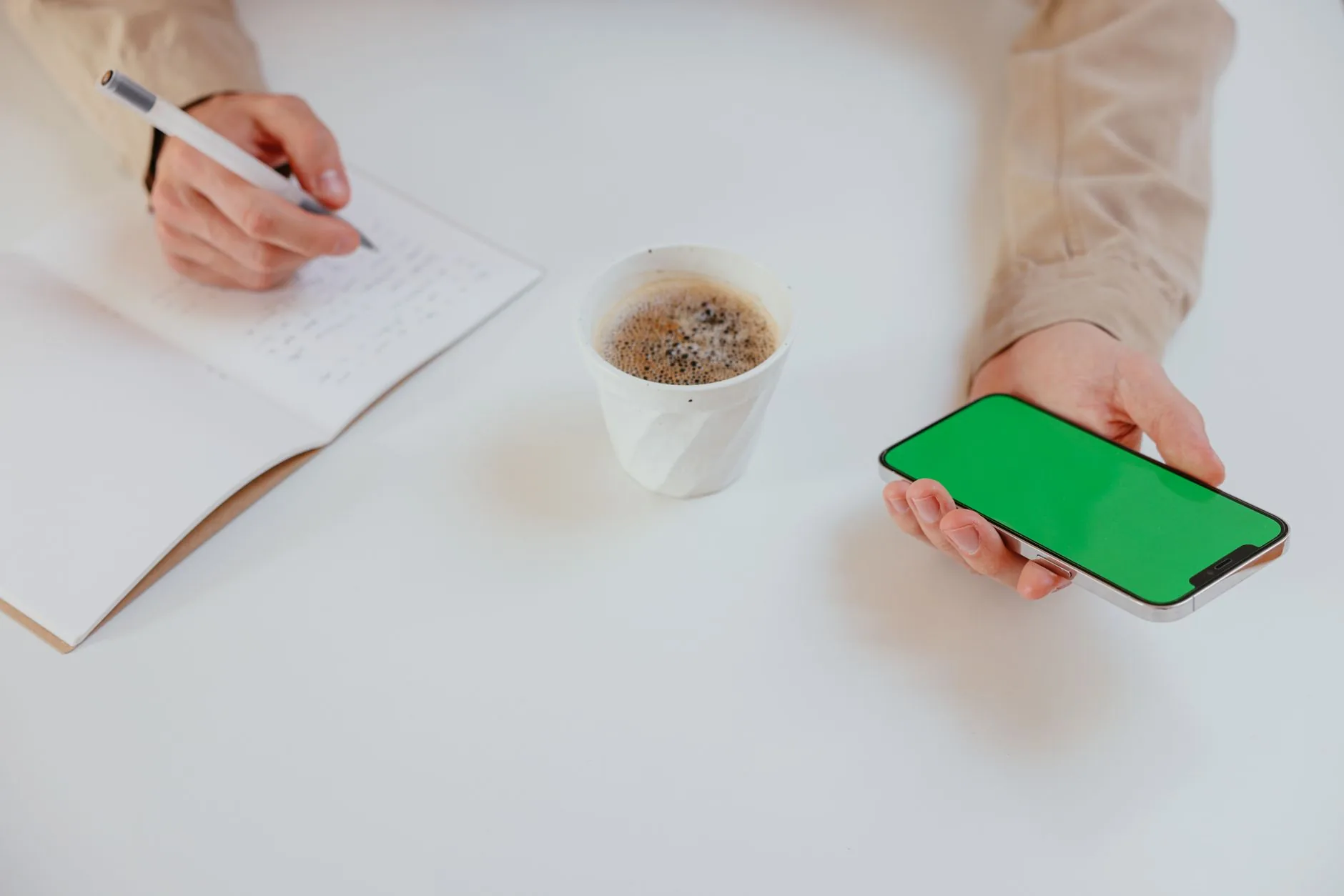 Close-up of hands holding a smartphone and writing notes with a coffee cup on a table.