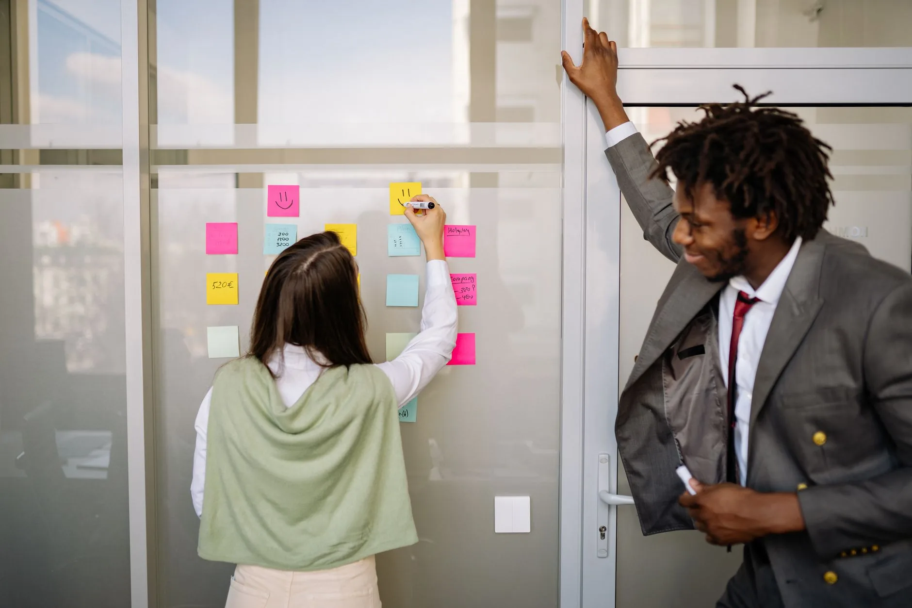 Two professionals brainstorming with sticky notes on a glass wall in a modern office setting.