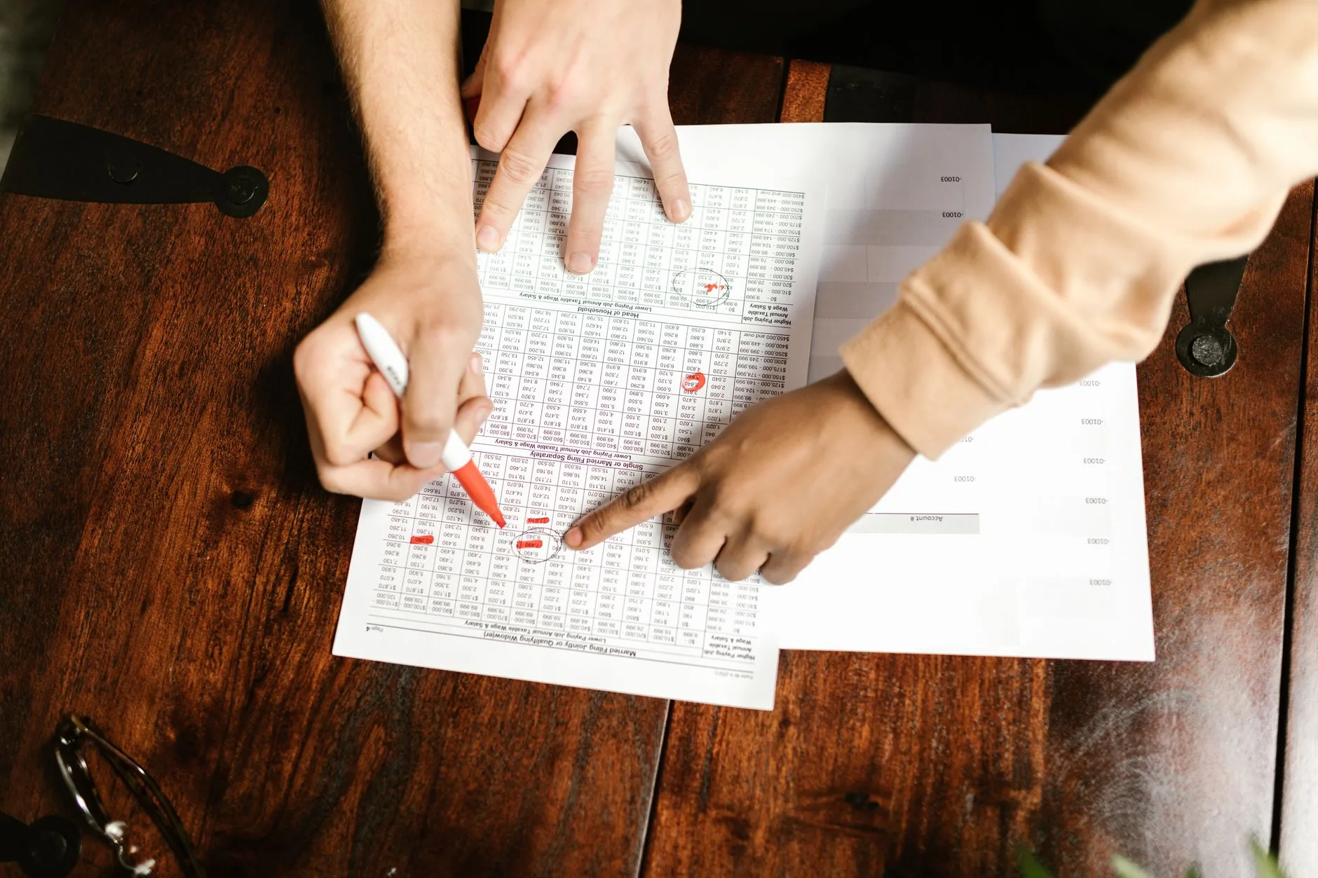 Two hands reviewing and marking financial documents with a red pen on a wooden table, top view.