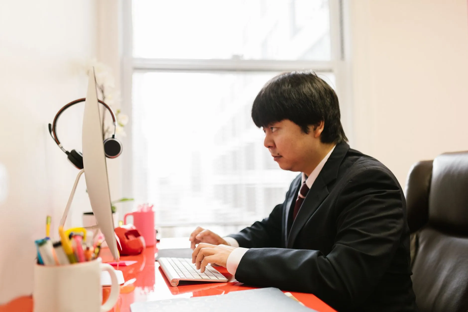 Asian businessman in a suit typing at a desktop computer in a modern office setting.