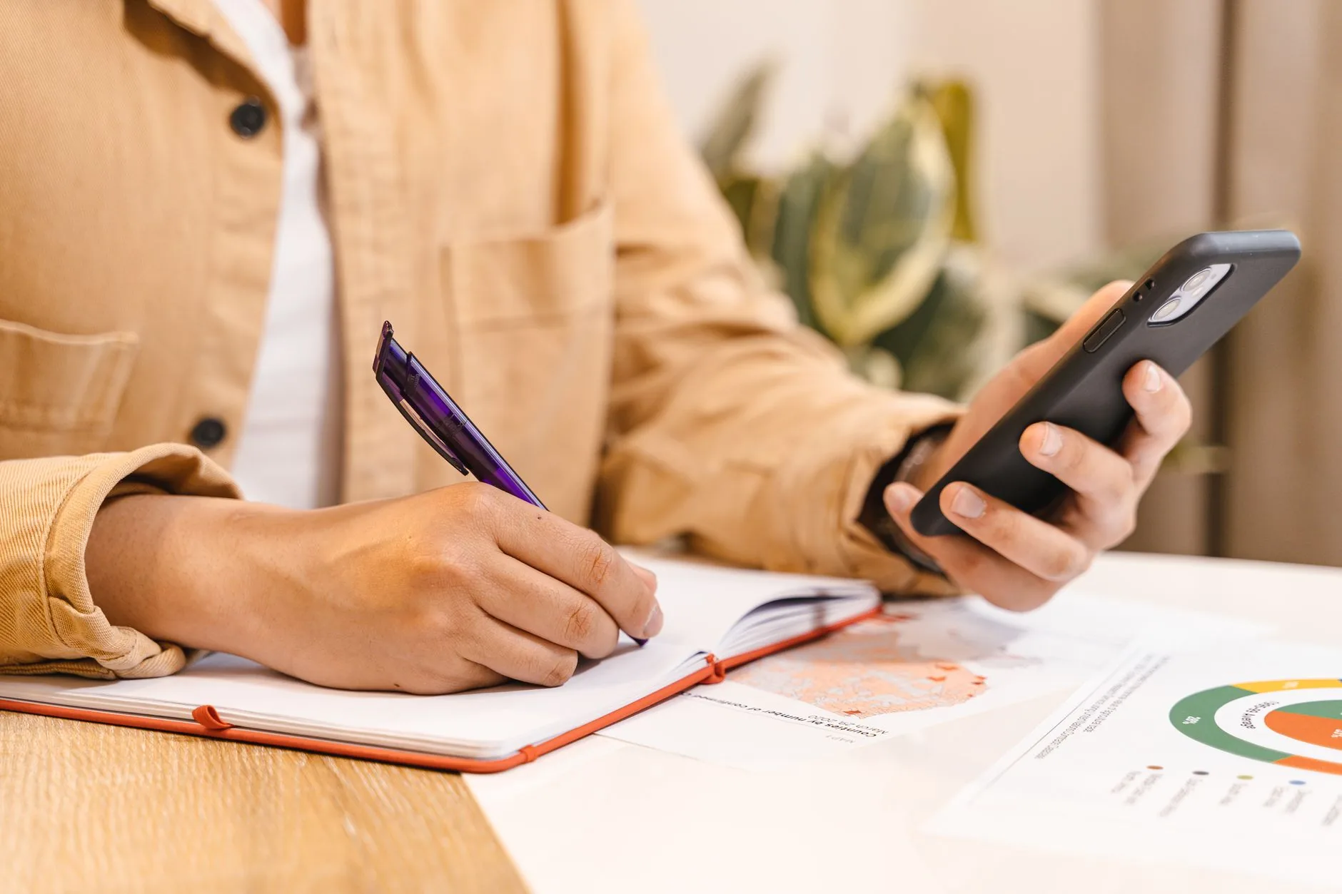 Close-up of hands holding a smartphone and writing in a notebook at a desk.