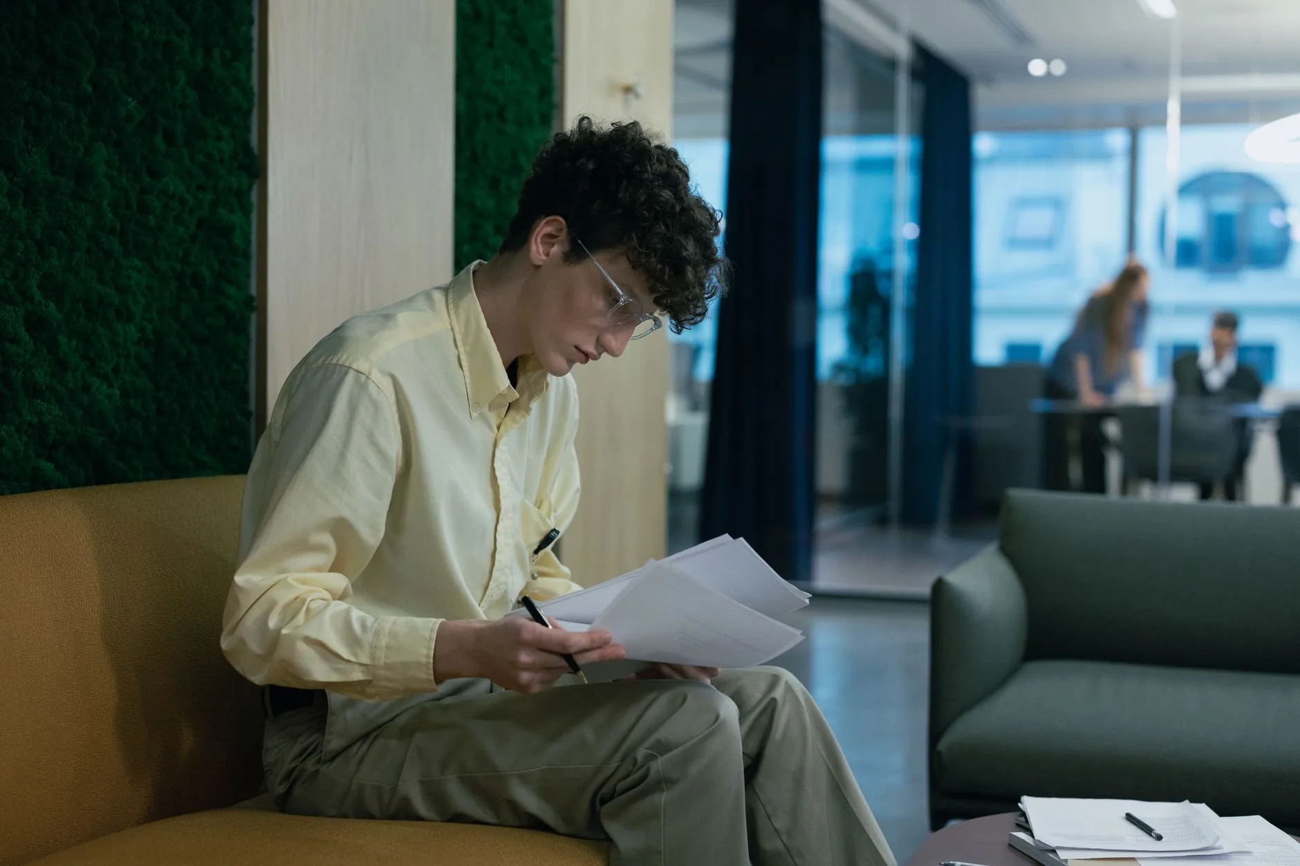 A focused young professional reading documents on a couch in a modern office environment.